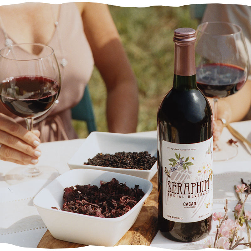 Friends around a table. Bottle of Seraphim Social Beverage, a non-alcoholic social wine alternative made from pure ingredients. Featured flavors in image: a bottle of Cacao with a poured clear glass.